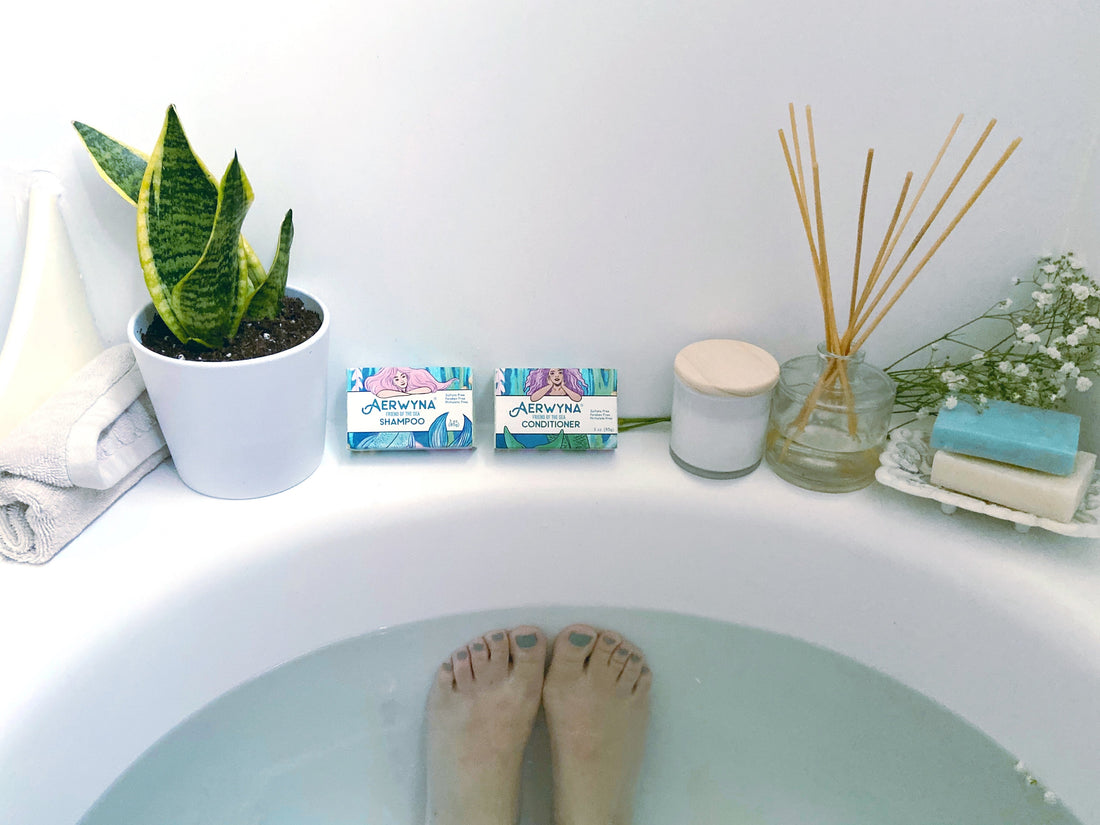 person relaxing in bath tub surrounded by Aerwyna shampoo and conditioner bars, a fragrant candle and snake plant.