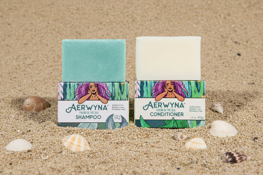 Aerwyna solid shampoo and conditioner bars with packaging resting in sand with sea shells